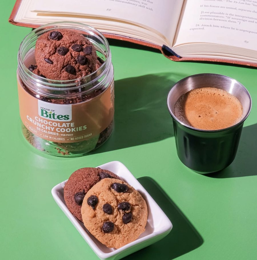 A House of Bites chocolate crunchy cookies jar beside an espresso, with two cookies on a small white plate and an open book behind.